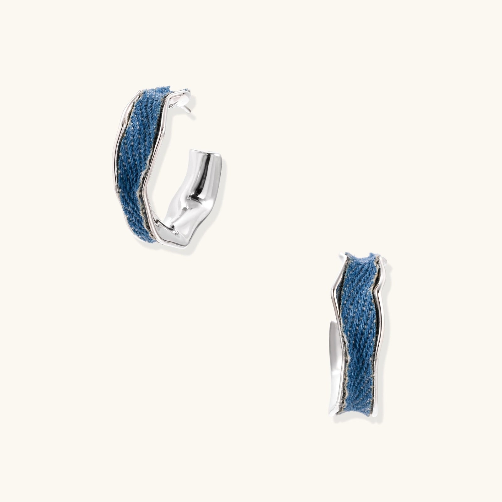 Silver huggie hoops with blue denim fabric inlay strip and polished white gold plated finish on cream background.
